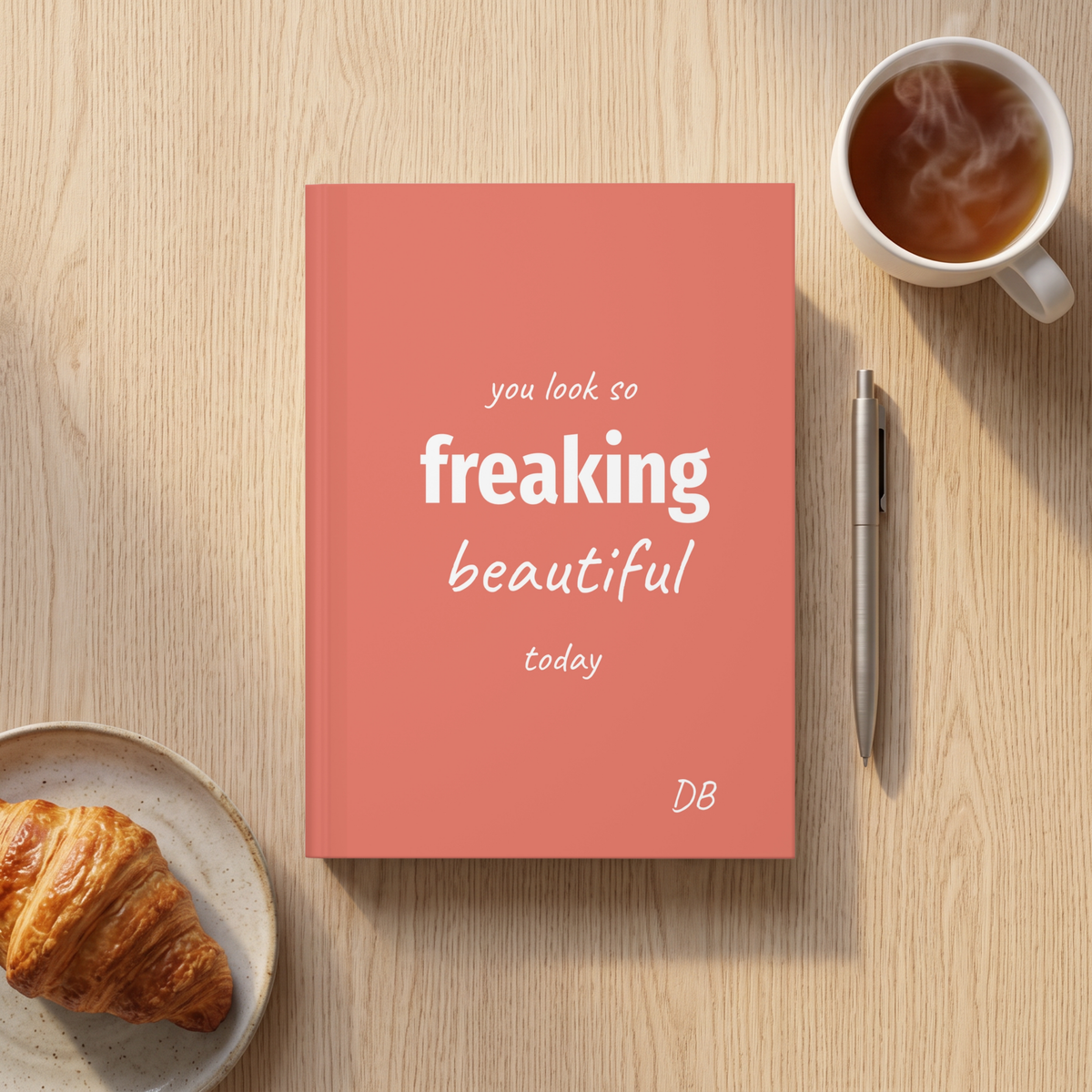 Coral Freaking Beautiful journal on a table next to a coffee and a croissant