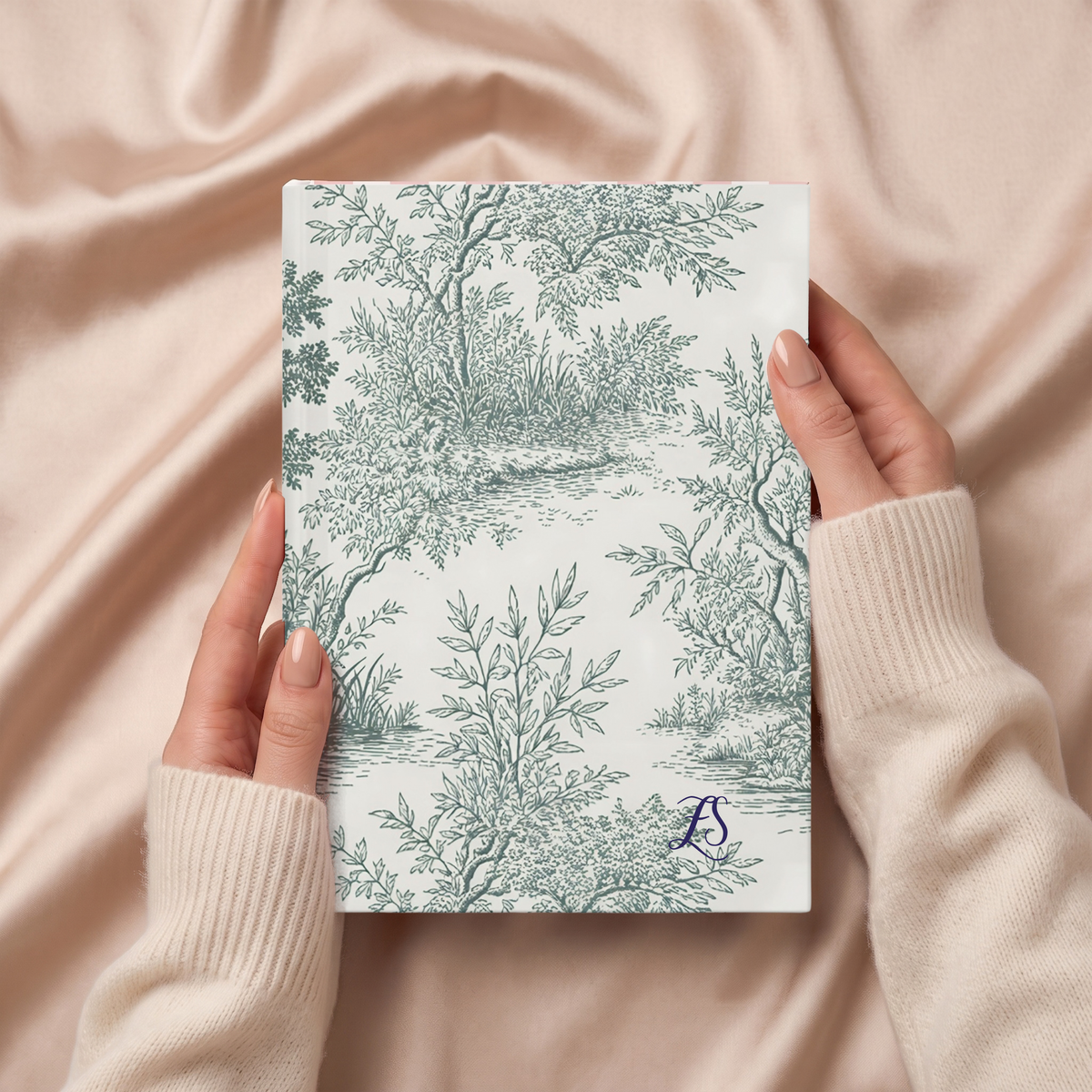 Hands holding the green Toile de Jouy forest journal on pink satin
