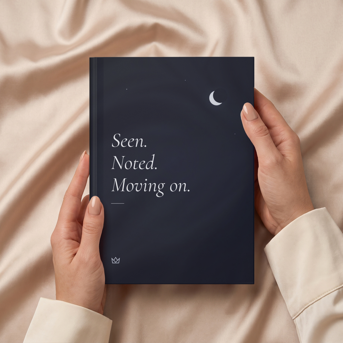 Hands holding the night-blue Seen. Noted. Moving on. journal over soft beige fabric
